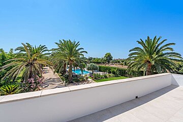 A sunny view from a white terrace shows lush gardens with numerous palm trees, a sparkling blue swimming pool, and a clear blue sky.