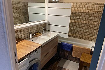 A compact bathroom features a double vanity sink, washing machine under a wooden counter, tiled walls, and a bathtub with a striped shower screen.
