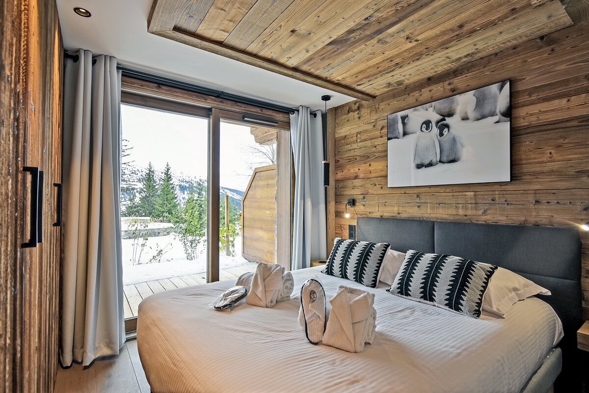Cozy ski chalet bedroom with wood walls and ceiling, a large window overlooking snowy mountains, a bed with towels, and penguin art.