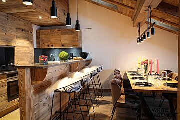 Cozy, rustic kitchen and dining space with wooden walls/ceiling, a bar with furry stools, and a dining table set with colorful candles.