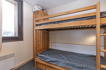 A room with wooden bunk beds and gray mattresses. A window offers a snowy mountain view. A wooden wardrobe and a wall heater are also visible.