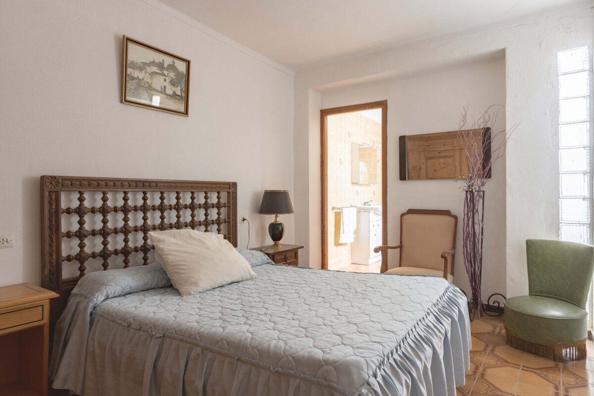 A cozy bedroom features a bed with an ornate wooden headboard and light blue bedding. A nightstand, lamp, two chairs, and a bathroom doorway are visible.