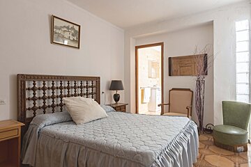 A cozy bedroom features a bed with an ornate wooden headboard and light blue bedding. A nightstand, lamp, two chairs, and a bathroom doorway are visible.