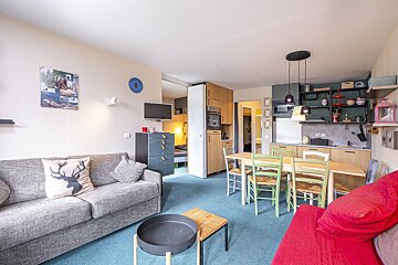 A cozy, open-plan studio apartment featuring a grey sofa, red couch, dining area, and kitchenette, all on blue carpet. A bed is visible in the background.