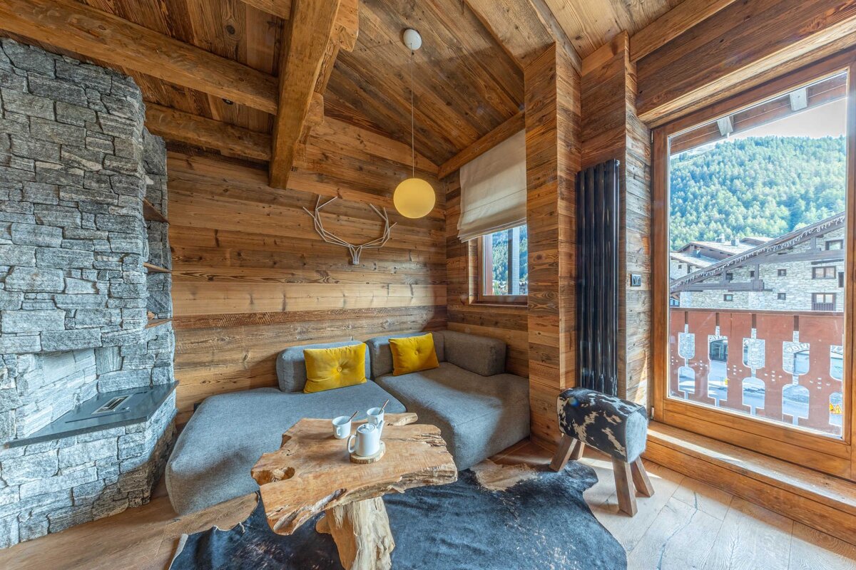 Cozy, rustic wooden chalet room with a stone fireplace, comfy sofa, and a stunning mountain view through a large window.