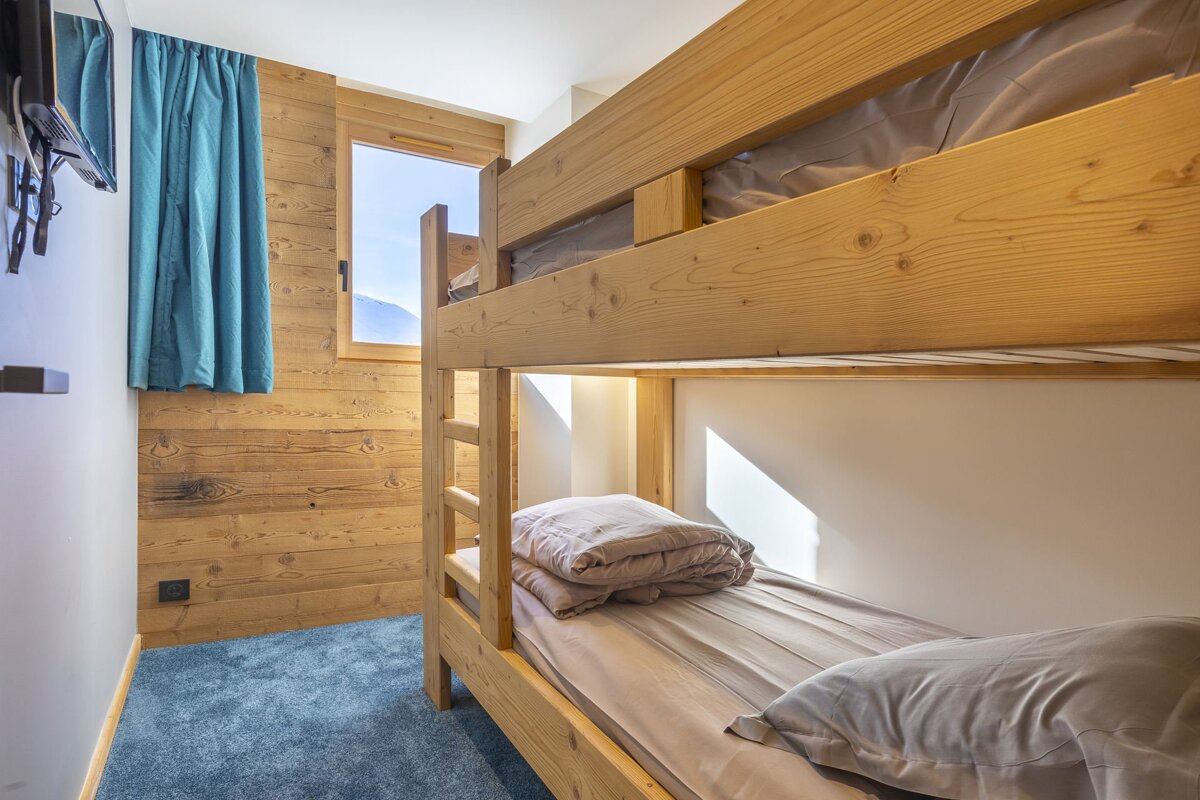 A cozy room features wooden bunk beds, a paneled wall, and a window revealing a mountain view. Blue carpet, a blue curtain, and a wall-mounted TV complete the space.