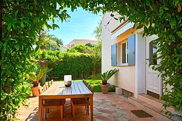 Sunny outdoor patio with a wooden dining set, bordered by lush green leaves, next to a house with blue shutters under a bright sky.