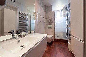 Modern bathroom with a white vanity, large mirror, toilet, frosted glass shower, dark wood floor, and chrome towel warmer.