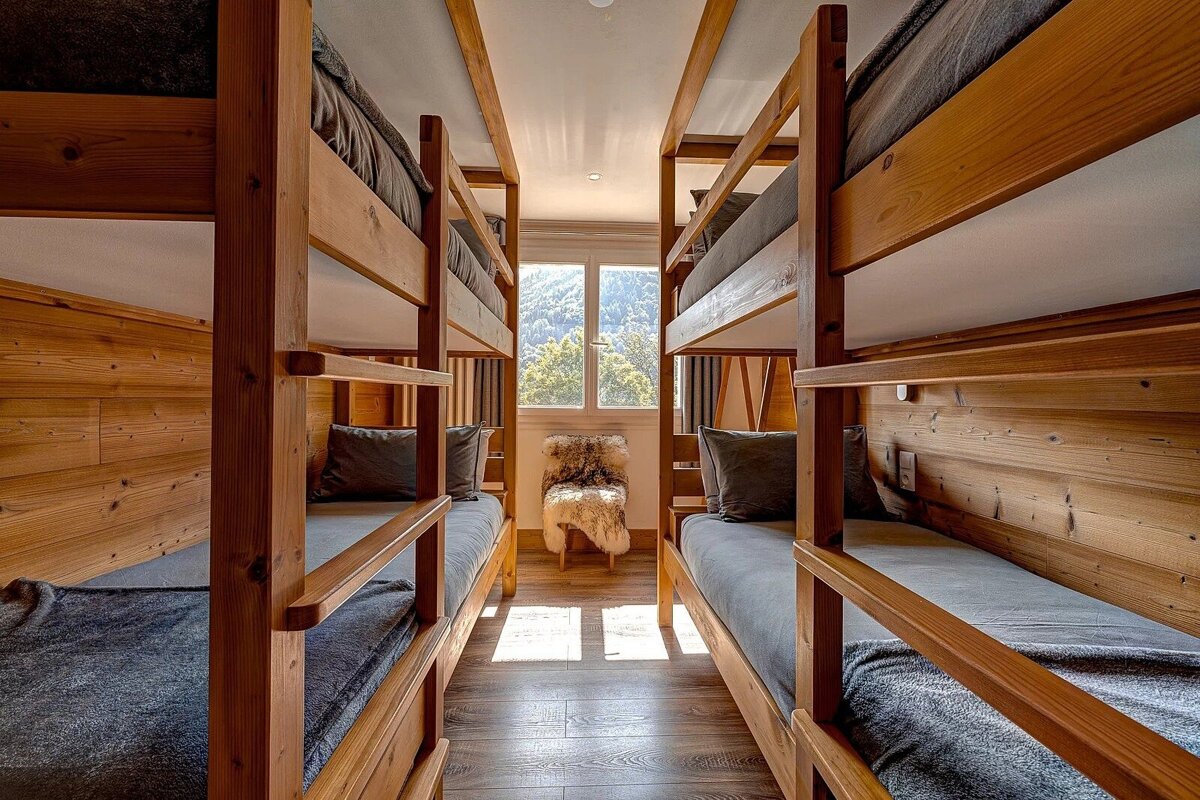 A rustic room features two wooden bunk beds facing each other, dark gray bedding, and a furry chair by a window overlooking mountains.
