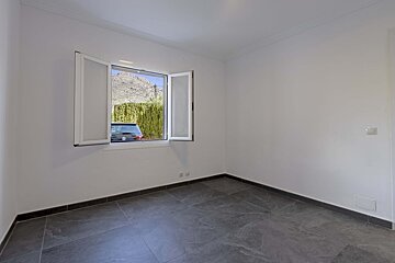 An empty room with white walls, dark tiled floor, and an open window revealing a car, green hedge, and a distant mountain view.