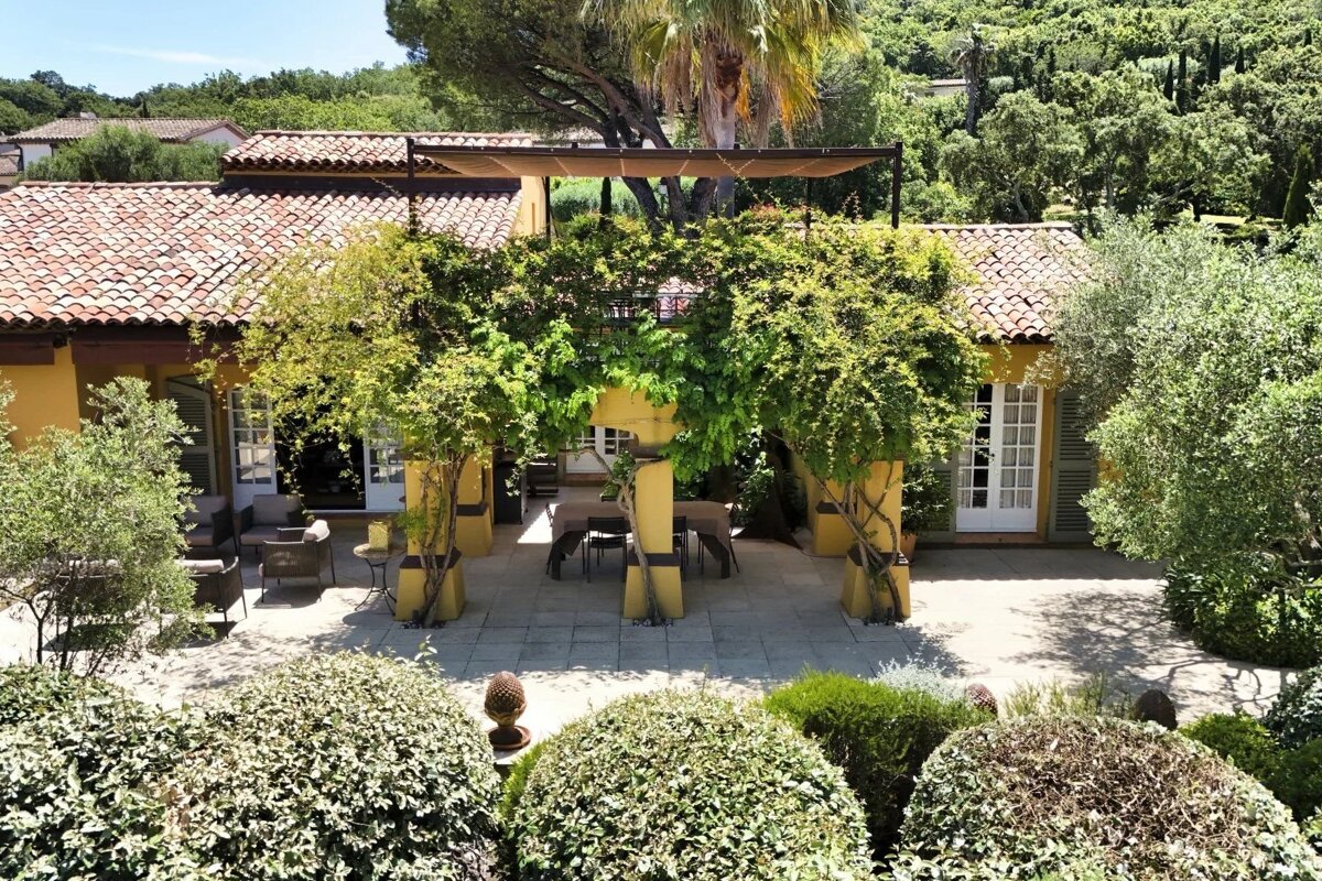 A beautiful yellow villa with a red tile roof features a vine-covered pergola over a stone patio with outdoor dining and lounge areas, surrounded by lush gardens.