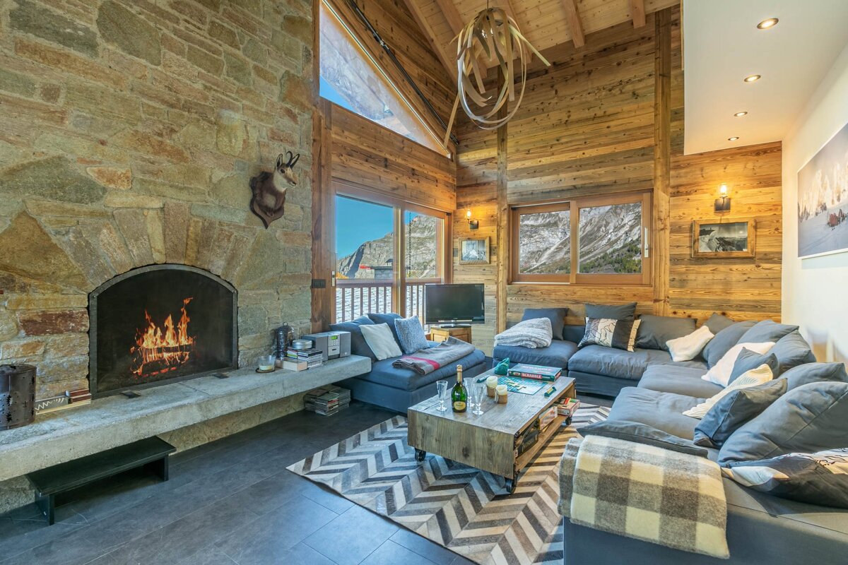Cozy wooden cabin living room with a large stone fireplace, comfortable sofas, and mountain views through the windows.