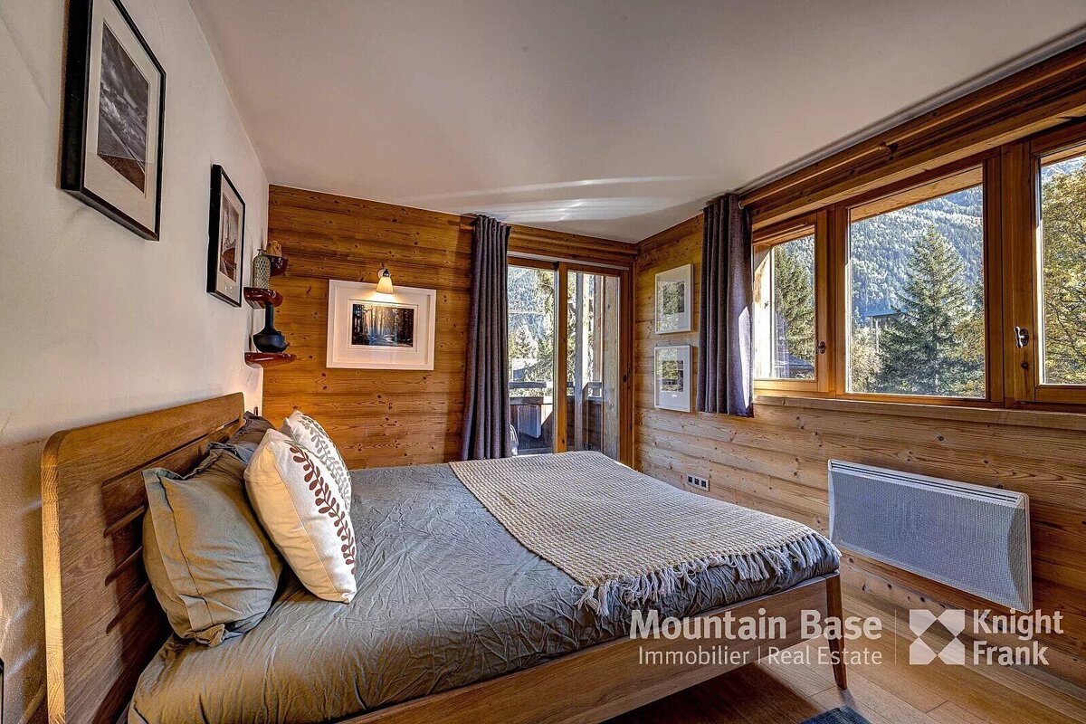A warm, wood-paneled bedroom with a comfy bed, balcony access, and large windows revealing a scenic mountain forest view.