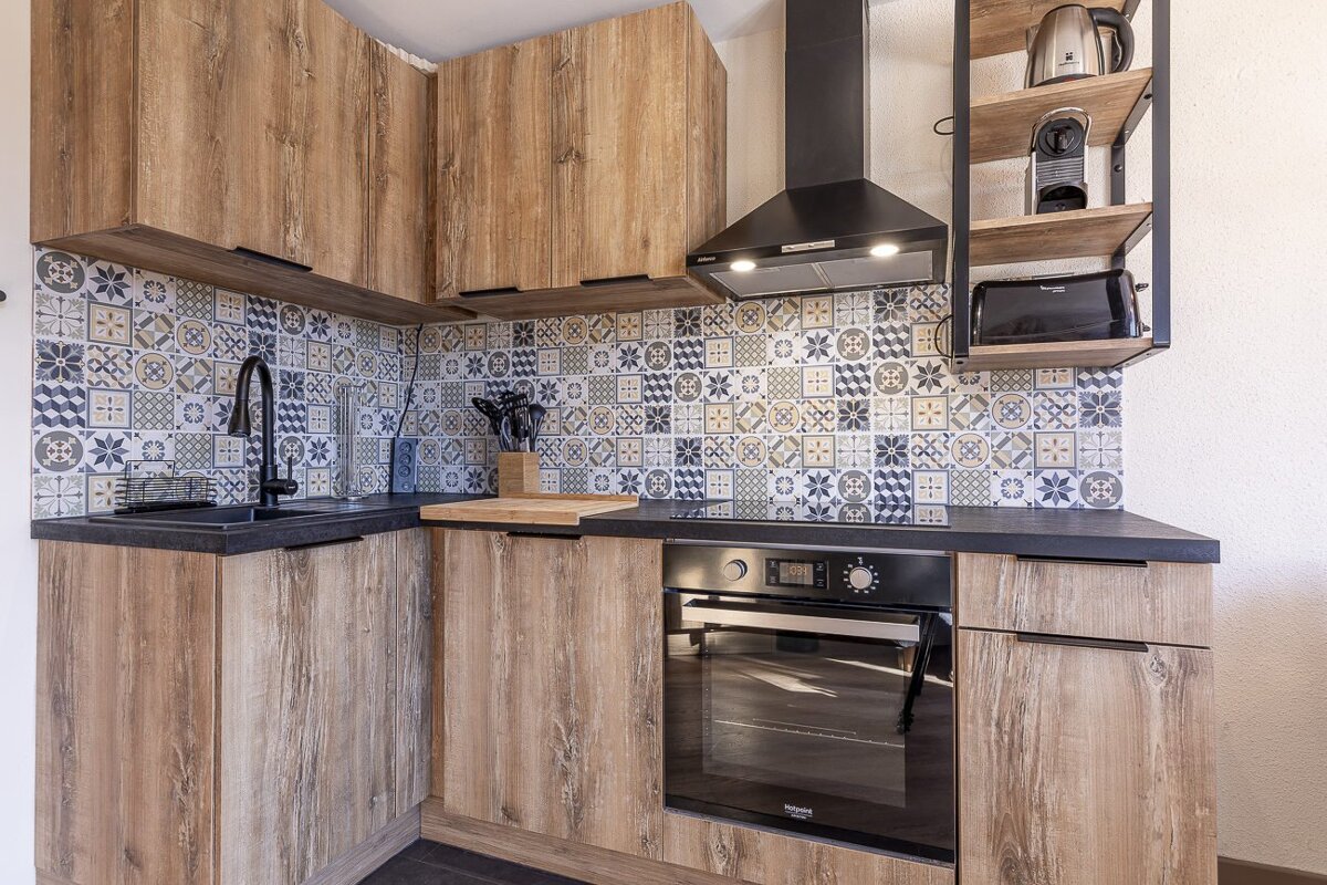 Compact, modern kitchen featuring wood-grain cabinets, black countertops, a colorful geometric tile backsplash, and built-in appliances.