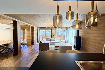 A modern, wood-paneled chalet interior with an open-plan living and dining area, featuring large windows, a sectional sofa, and stylish pendant lights.