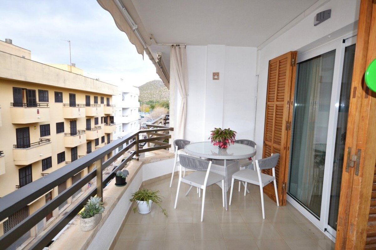 An apartment balcony featuring a glass-topped dining table, four chairs, potted plants, and views of surrounding buildings.