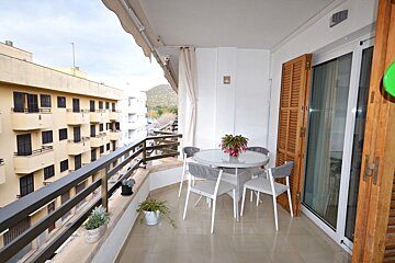 An apartment balcony featuring a glass-topped dining table, four chairs, potted plants, and views of surrounding buildings.