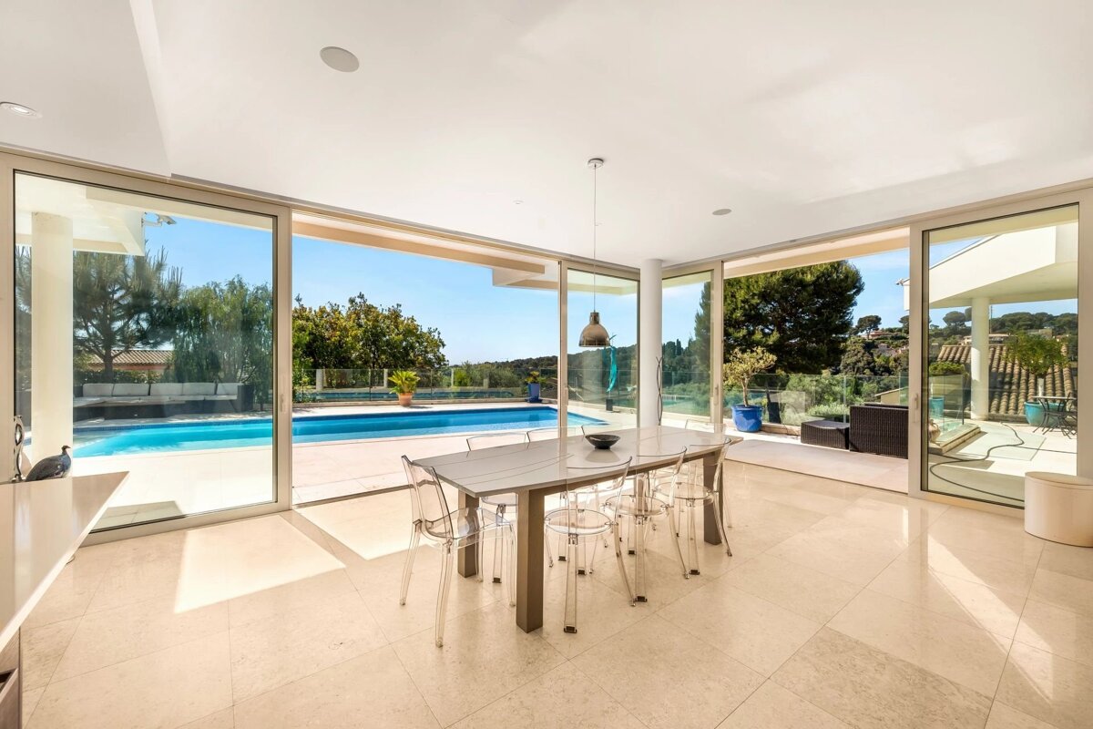 A modern dining room with a table and clear chairs, overlooking a swimming pool and patio through large glass sliding doors.