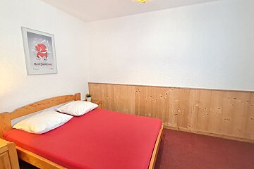 A bedroom featuring a wooden bed with a vibrant red sheet, white pillows, wood paneling, and a ski-themed poster on the wall.
