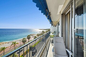 Sunny balcony with a blue awning overlooks a vibrant blue sea, pebble beach, palm trees, and a coastal road with traffic.