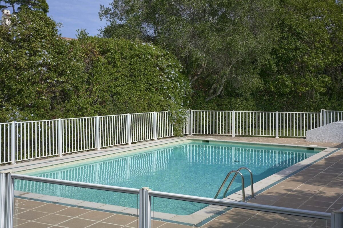 A fenced outdoor swimming pool with clear blue water, surrounded by a tiled patio and lush green foliage under a bright sky.