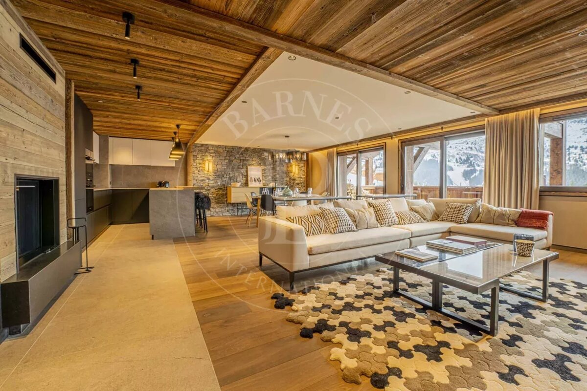 Modern chalet apartment with an open living space, wooden ceilings, stone accent wall, sleek kitchen, and large windows revealing snowy mountain views.