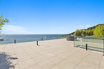 Spacious modern terrace with beige tiles and glass railings, offering a panoramic view of the blue ocean under a clear sky.