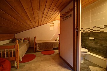 A cozy attic bedroom with two single beds, a sloped wooden ceiling, and light wood floors with red rugs. An open door shows a modern bathroom with a toilet.