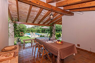 A table and chairs under a wooden roof with a pool in the background