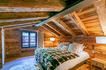 A cozy, rustic bedroom with warm wooden walls and a slanted ceiling featuring exposed beams. A bed with a green and white faux fur throw sits by a window.