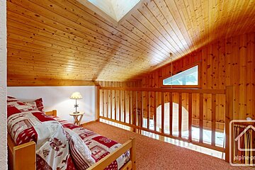 A rustic wood-paneled loft bedroom featuring a bed with patterned bedding, a lamp, and a triangular window. A wooden railing overlooks a lower level.