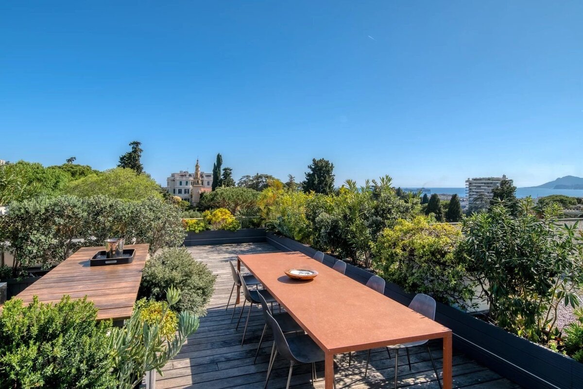 Sunny rooftop terrace with wooden decks, dining tables, lush plants, and panoramic views of a distant city, sea, and clear blue sky.