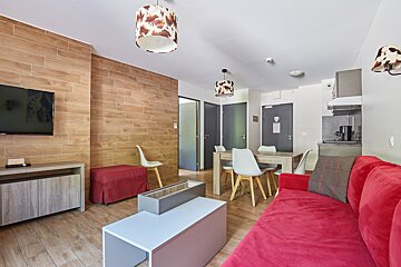 A modern living room/dining area with a red couch, wooden accents, and a small kitchen. Features cow print light fixtures and light wood flooring.