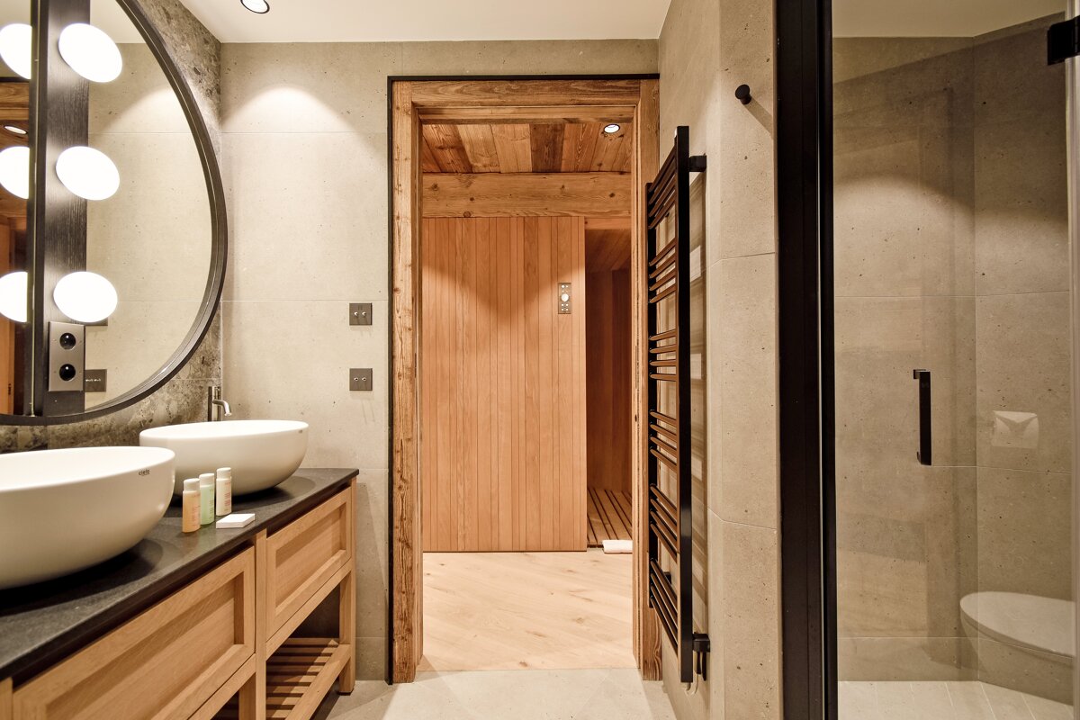 A modern bathroom features double sinks, a lighted mirror, tiled walls, and a wooden sauna visible through a doorway. A shower and towel warmer are also present.