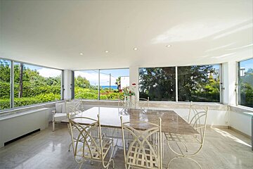 Sunlit dining room with extensive windows showcasing a panoramic view of vibrant green foliage and the sparkling blue sea beyond.
