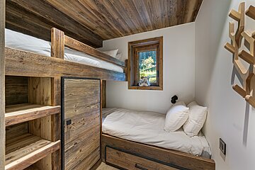 Cozy rustic bedroom with wooden bunk bed, a single bed below, and a window with a green view. Wood-paneled ceiling and built-in storage.