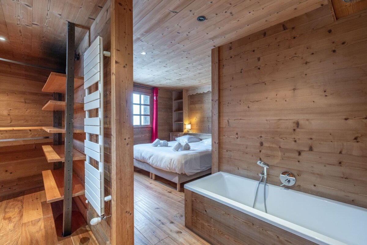 A rustic, all-wood interior featuring a bedroom with a cozy bed, red curtains, and window, alongside an open bathtub area. Wooden shelves and a white radiator are visible.