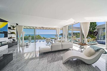 Spacious, bright living room with white furniture, marble floor, and stunning ocean views through floor-to-ceiling glass walls.