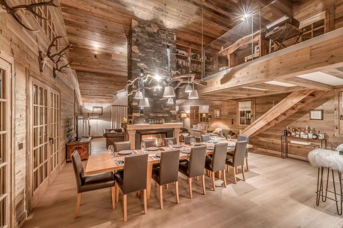 Luxurious rustic chalet interior with a grand dining table, central stone fireplace, warm wood paneling, a mezzanine, and inviting lighting.