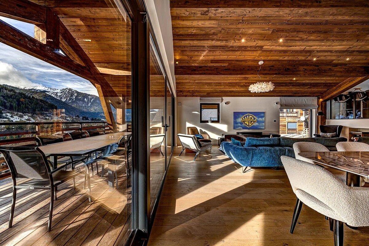 Modern alpine chalet interior featuring wood paneling, a blue sofa, and large glass doors opening to scenic mountain views.