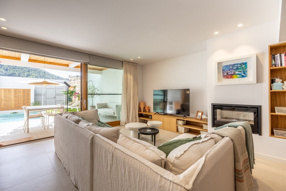 Modern living room with a large sofa, TV, and fireplace. Sliding doors open to a patio with a pool, outdoor dining, and mountain views.