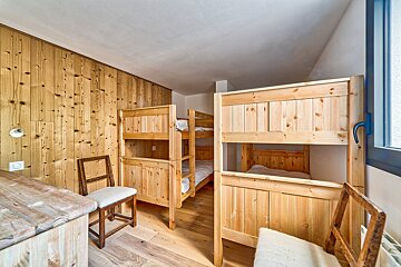 A rustic room with several wooden bunk beds, wood-paneled walls, and a wooden floor. A desk and chairs provide a cozy, shared sleeping space.