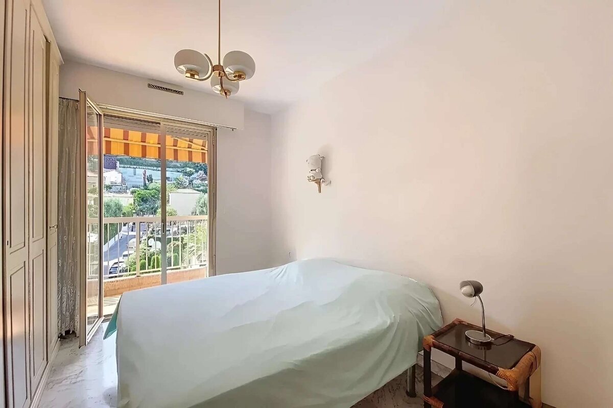 A simple, bright bedroom featuring a bed, a large balcony with a scenic outdoor view, a wardrobe, and a mid-century style chandelier.
