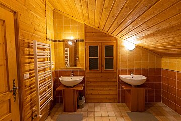 Rustic, attic-style bathroom with slanted wooden ceiling, wood-paneled walls, and two modern white sinks. Brown and yellow tiled walls add color.