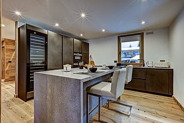 A modern kitchen features a large gray island with bar stools, dark cabinetry, wood floors, and a wine cooler. Breakfast items are laid out, and a window reveals a snowy outdoor scene.
