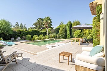 Sunlit patio with a pool, outdoor seating, a wooden table with an umbrella, and lush green landscaping under a clear sky.