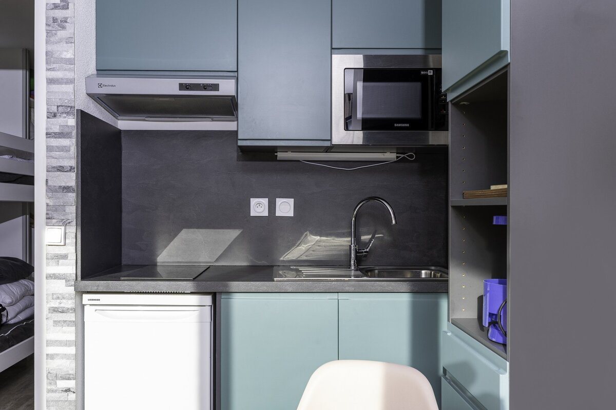 A modern compact kitchen with light blue cabinets, grey countertop, sink, hob, microwave, fridge, and range hood. Bunk bed visible on the left.