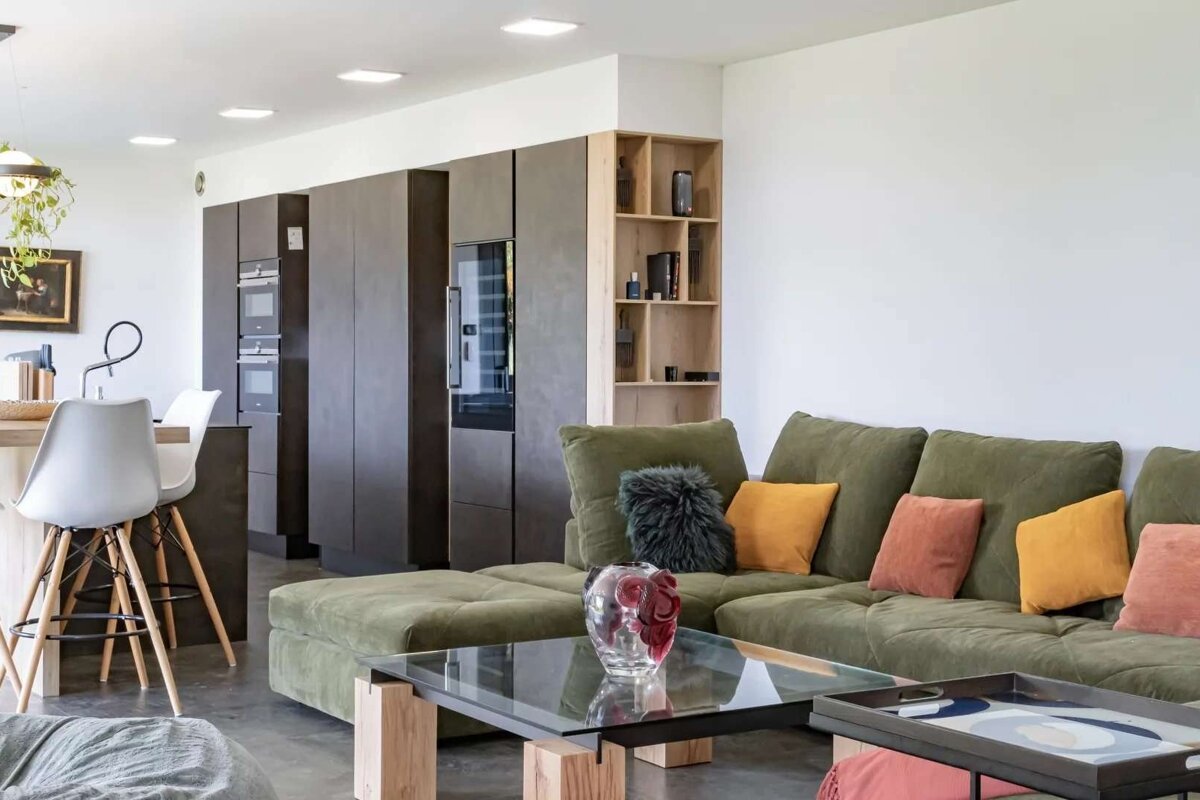 Spacious, modern open-plan living room and kitchen. Features an olive green sofa with vibrant cushions, dark kitchen cabinetry, and white walls.