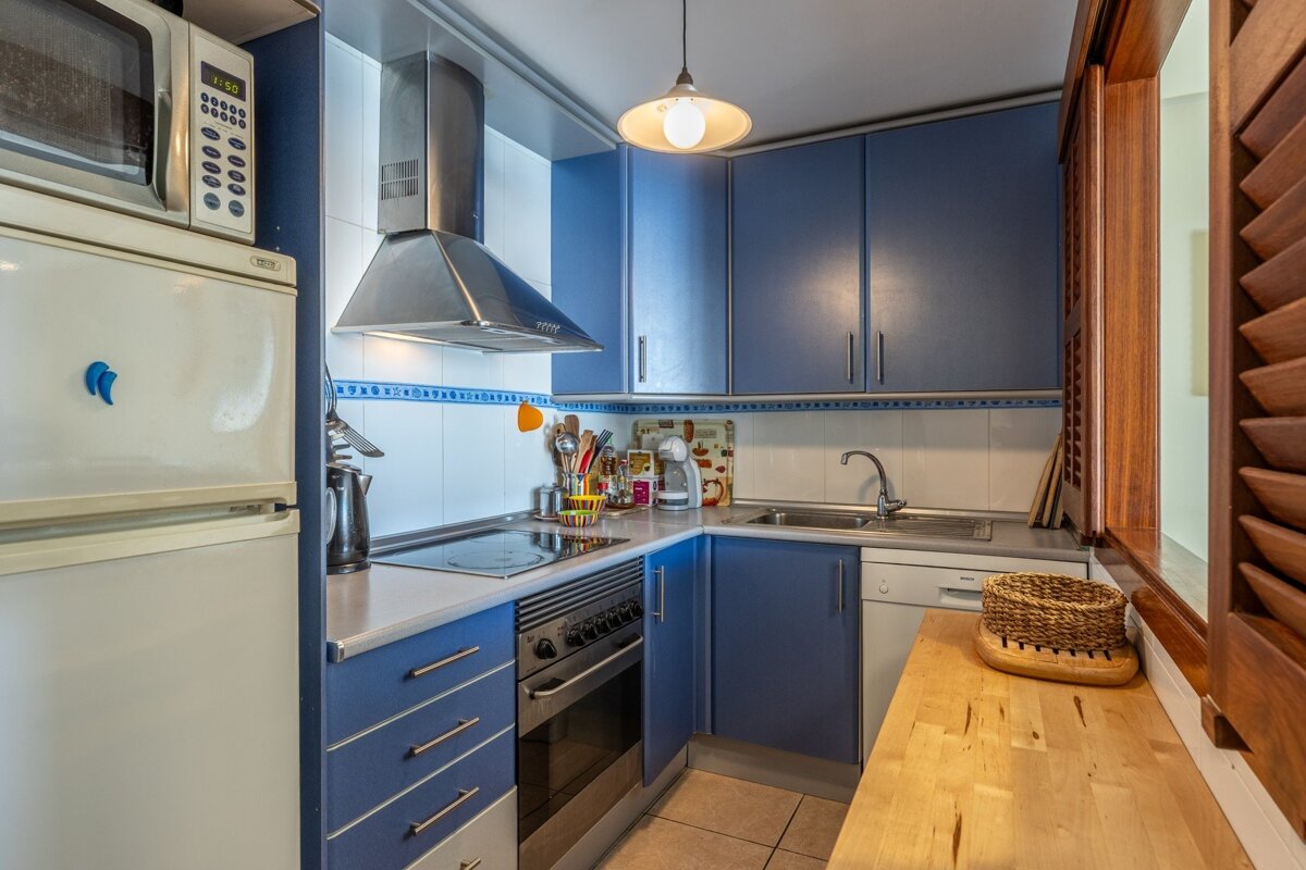 A compact blue and white kitchen with modern appliances like a fridge, microwave, and stove. Wooden shutter and countertop add a cozy feel.