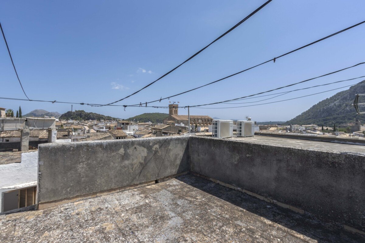 Rooftop view over a town with red-tiled roofs, a church, and mountains under a clear sky. Power lines crisscross, and AC units sit on the roof.